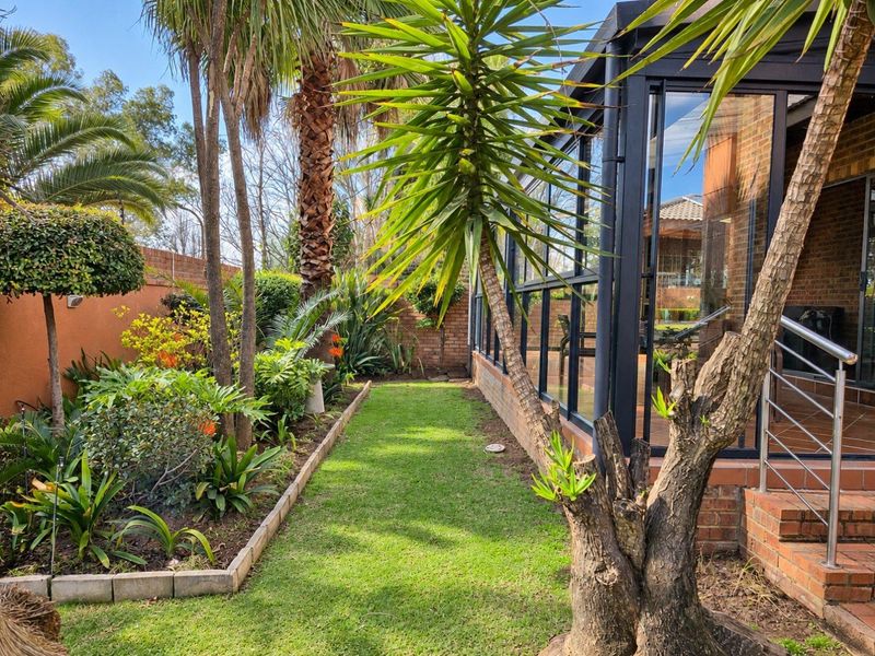 A well-maintained rear garden featuring a glass-enclosed patio, brick boundary walls, and established landscaping with mature trees and shrubs.