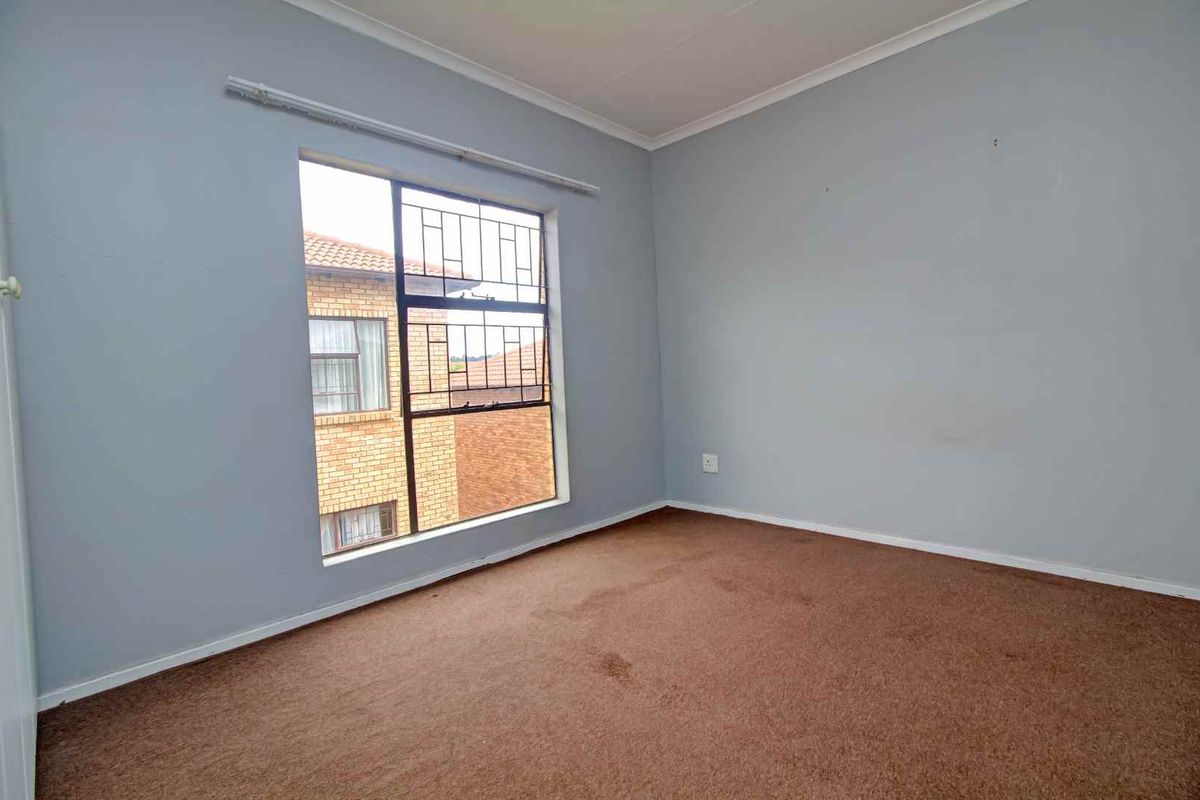 A vacant bedroom featuring neutral grey walls, brown carpeting, and a large window with security bars.