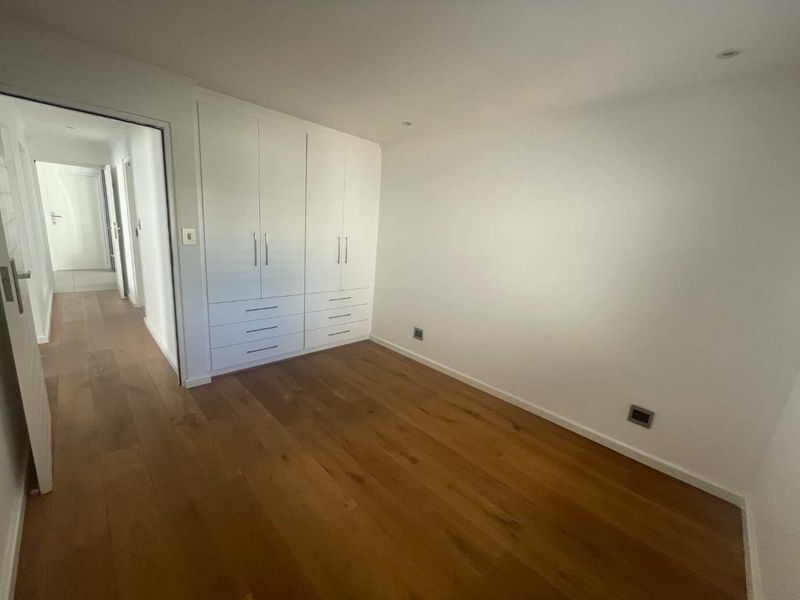 A bright, empty bedroom featuring polished hardwood flooring, neutral walls, and ample built-in cupboard storage with integrated drawers.