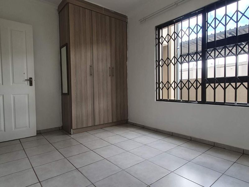 A clean, empty bedroom featuring neutral tiled flooring, a built-in wooden wardrobe, and a window fitted with a security gate.