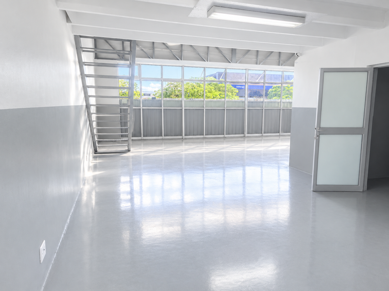 A bright, open-plan commercial or industrial space featuring polished epoxy flooring, a metal staircase, and expansive windows providing ample natural light.