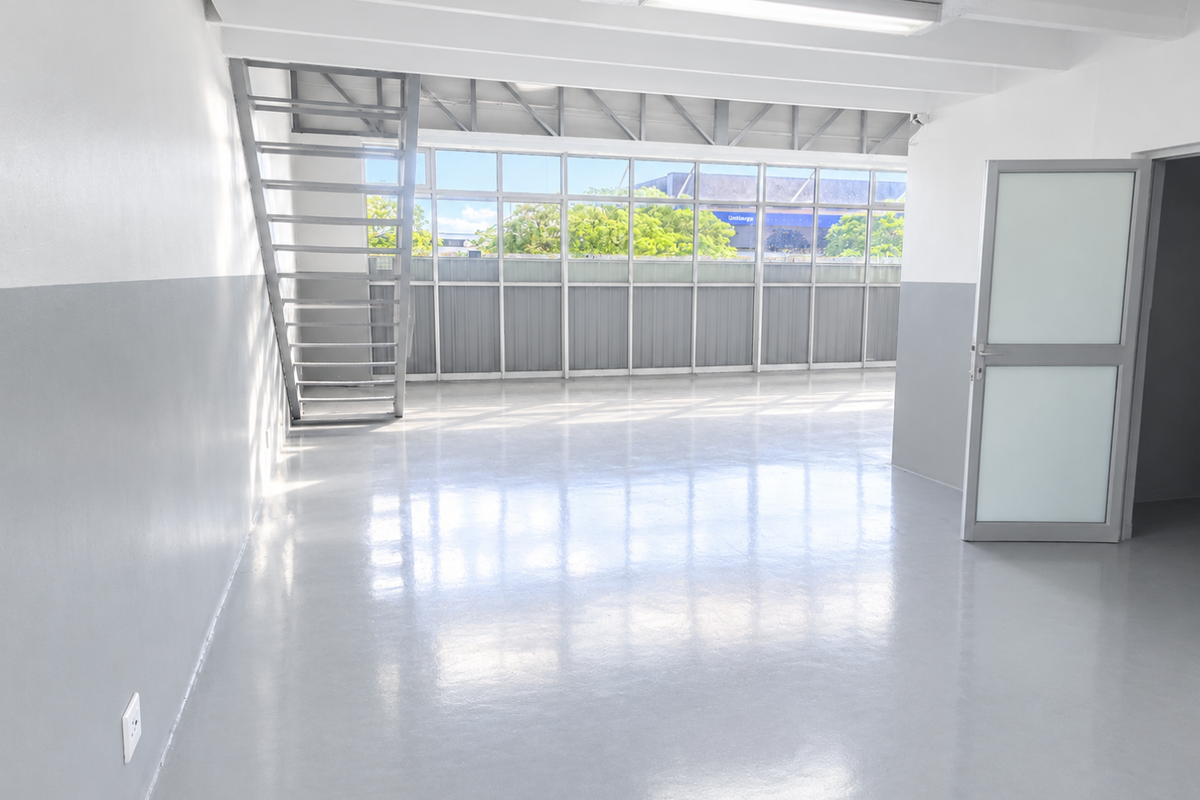 A bright, open-plan commercial or industrial space featuring polished epoxy flooring, a metal staircase, and expansive windows providing ample natural light.