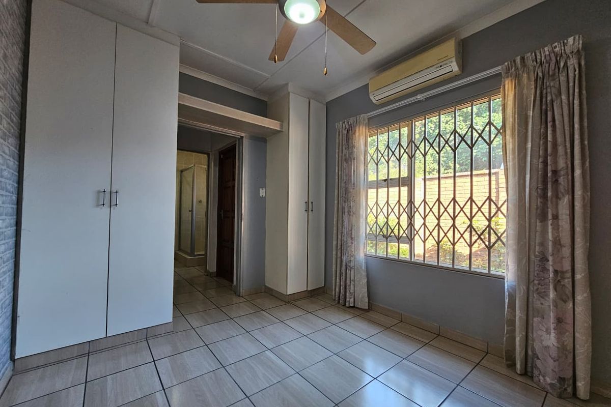 A bedroom featuring built-in cupboards, tiled flooring, a ceiling fan, and a window fitted with security bars.