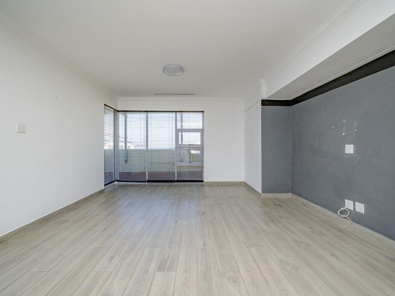 A bright, empty living room featuring light-toned laminate flooring, a grey accent wall, and large windows fitted with venetian blinds.