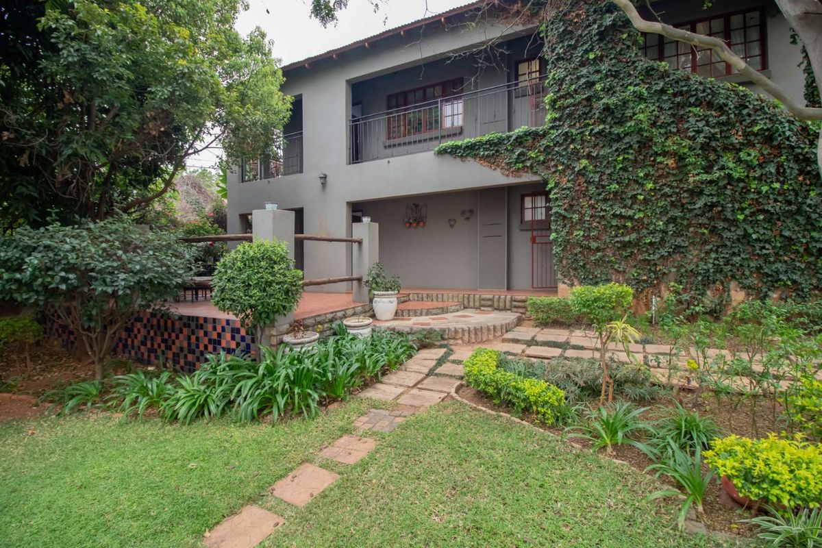 Rear view of a two-story guest house featuring a ground-level patio, upper balcony, and established garden with stone pathways.