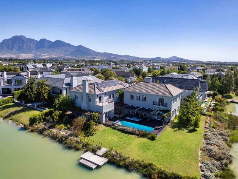 Aerial view of a luxury waterfront property featuring a swimming pool, wooden deck, pergola, private jetty, and solar panels with mountain views.