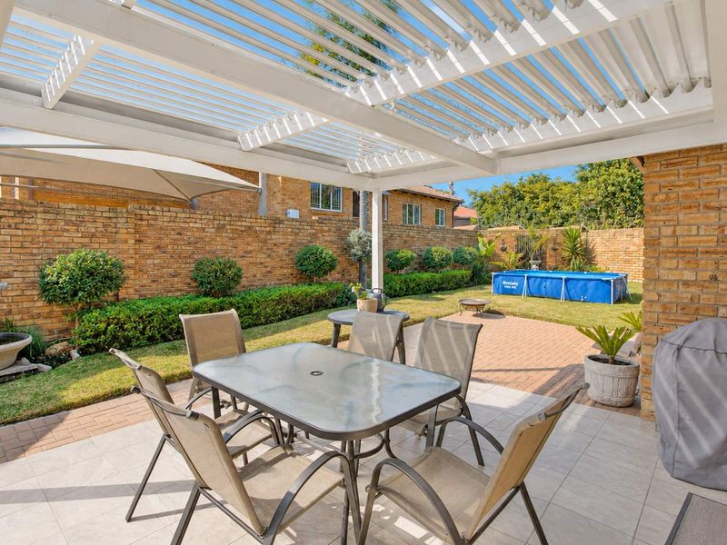 A well-maintained outdoor patio featuring a white pergola, paved flooring, and a spacious garden area with a brick boundary wall.
