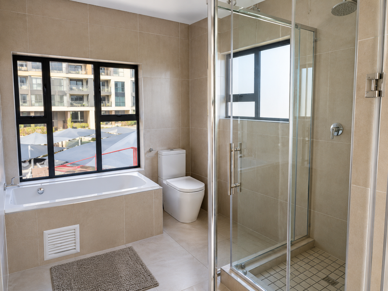 Modern bathroom featuring a built-in bathtub, separate glass-enclosed shower, and neutral floor-to-ceiling tiling with large windows providing natural light.