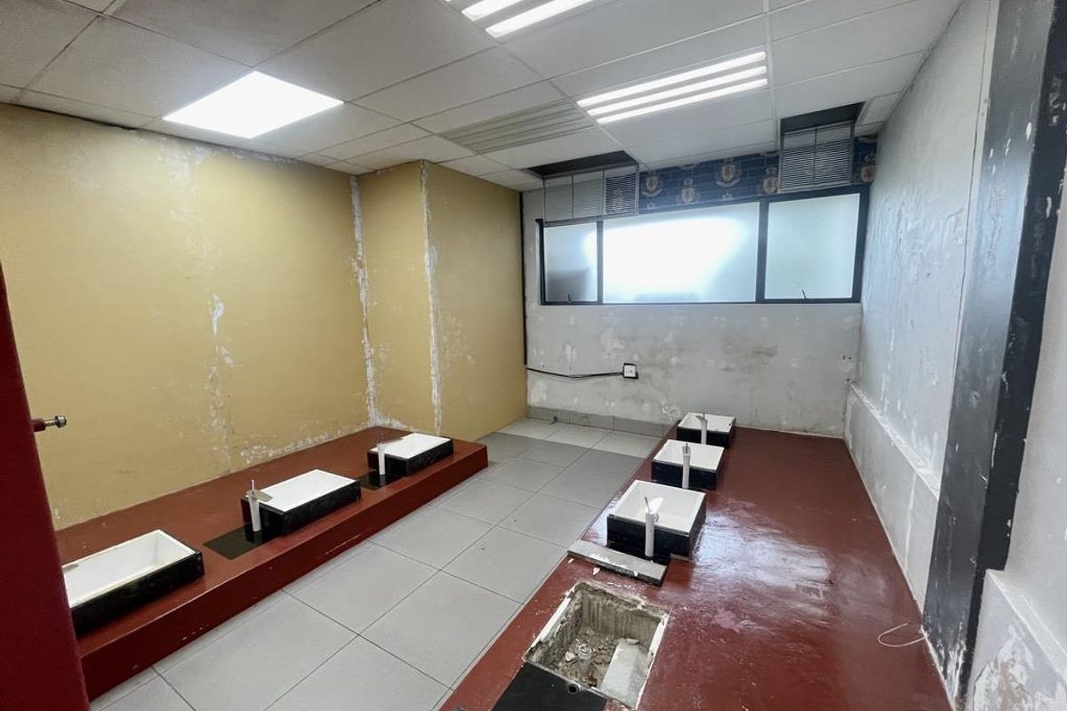 A vacant commercial washroom area featuring multiple built-in basins, tiled flooring, and suspended ceiling panels, currently requiring significant renovation and cosmetic repairs.