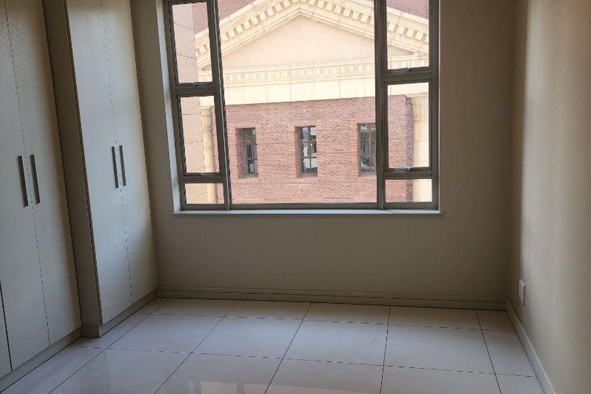 A bright, empty bedroom featuring neutral tiled flooring, built-in cupboards, and large aluminium-framed windows providing natural light.