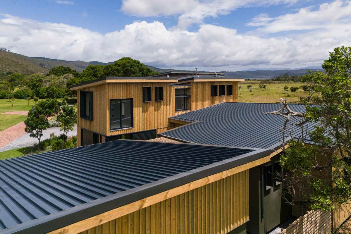 An elevated view of a modern two-story home featuring timber cladding, dark metal roofing, and large windows set within a scenic landscape.