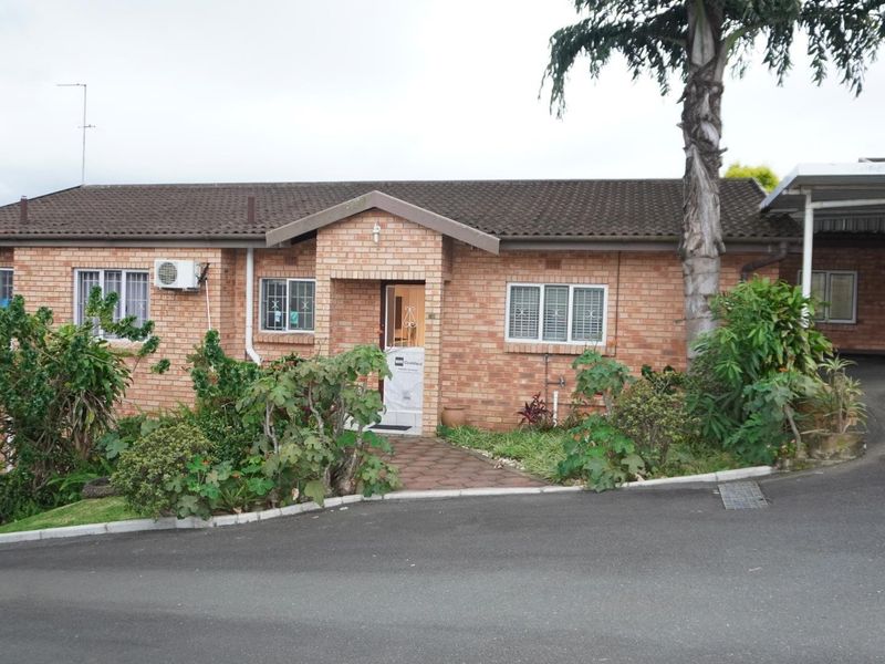 Front elevation of a face brick townhouse featuring a tiled roof, attached carport, and paved entrance walkway with established garden beds.