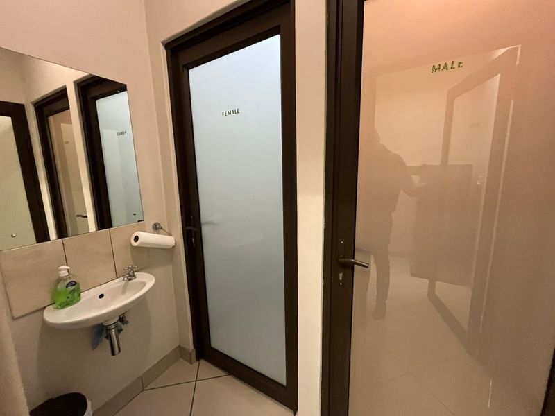 A clean office restroom area featuring a wall-mounted basin, tiled splashback, and separate frosted glass doors for male and female facilities.