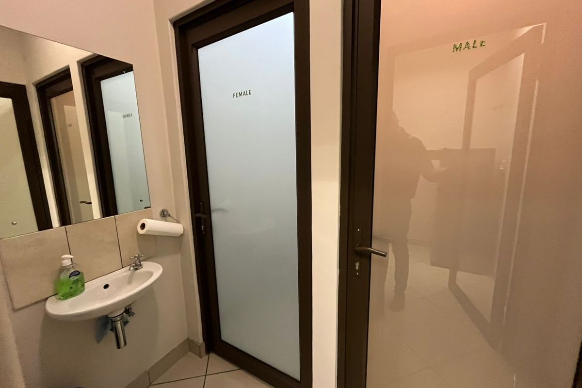 A clean office restroom area featuring a wall-mounted basin, tiled splashback, and separate frosted glass doors for male and female facilities.