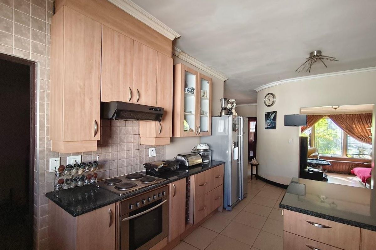 A functional kitchen featuring light wood cabinetry, granite countertops, a built-in oven with hob, and tiled finishes, flowing into an adjacent living area.