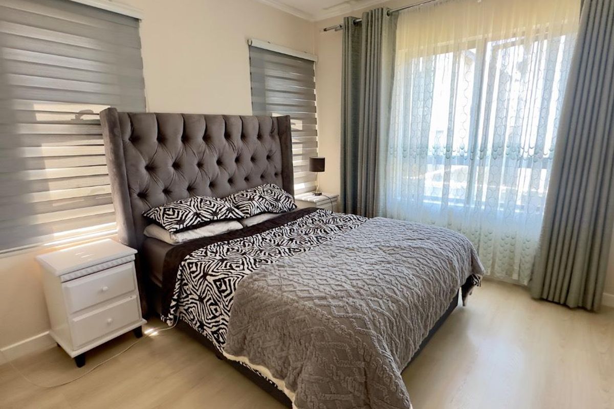 A well-maintained bedroom featuring light-toned laminate flooring, neutral wall finishes, and dual window treatments including roller blinds and floor-to-ceiling curtains.