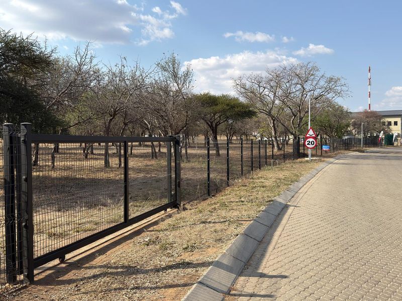 A view of the vacant commercial land featuring secure metal mesh fencing, a sliding gate, and paved road access with concrete curbing.