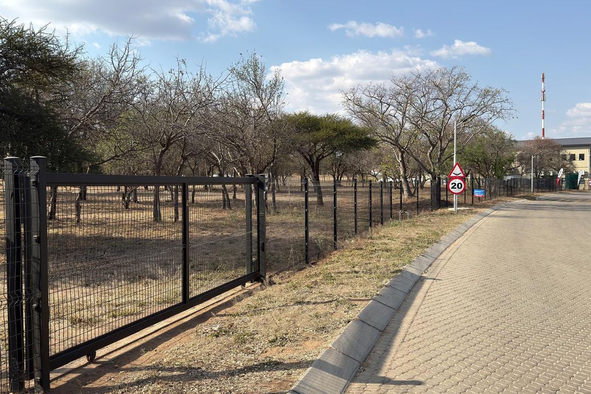 A view of the vacant commercial land featuring secure metal mesh fencing, a sliding gate, and paved road access with concrete curbing.