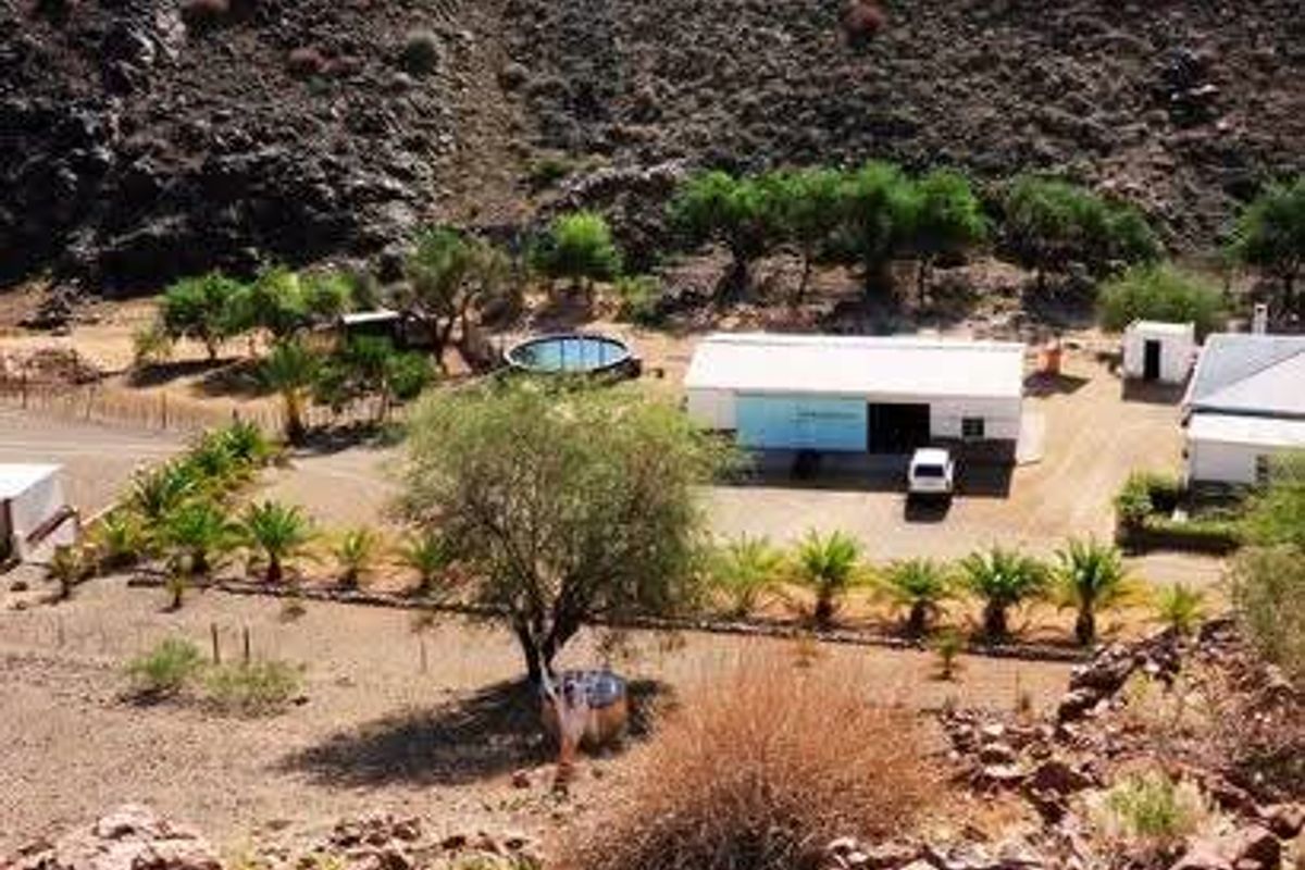 Aerial view of the farm property featuring a main building, outbuildings, a circular pool, and landscaped garden areas set against a rocky hillside.