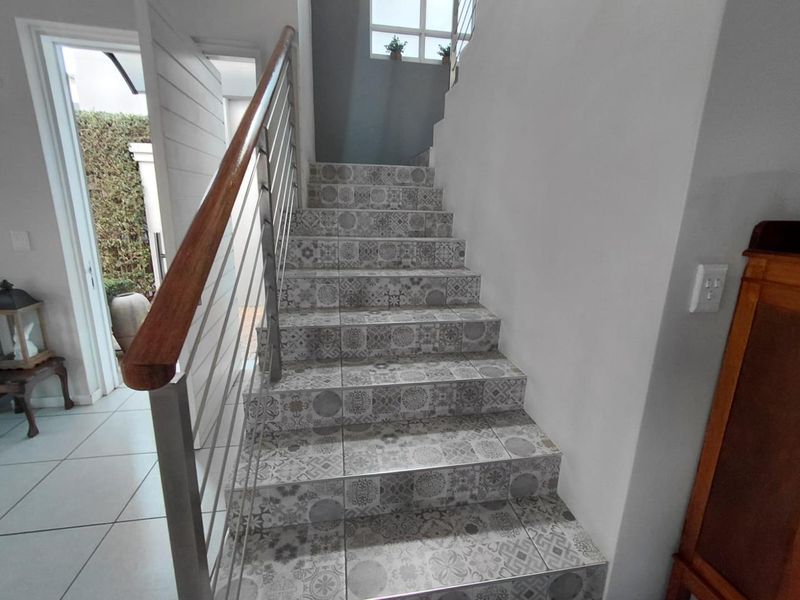 A bright hallway featuring a staircase with decorative patterned tiles, a wooden handrail, and a modern stainless steel balustrade.