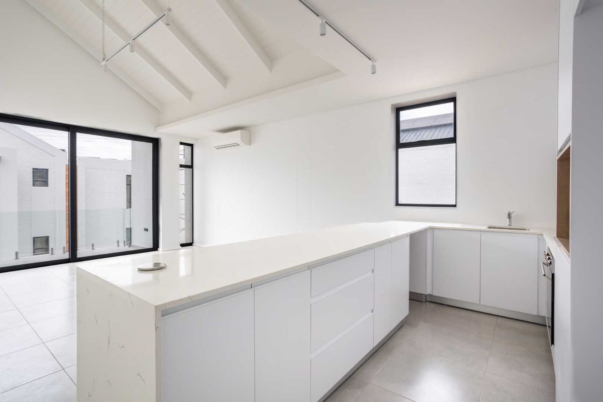 Modern, minimalist kitchen featuring a large stone island, sleek white cabinetry, built-in oven, and vaulted ceilings with track lighting, opening to an outdoor area.