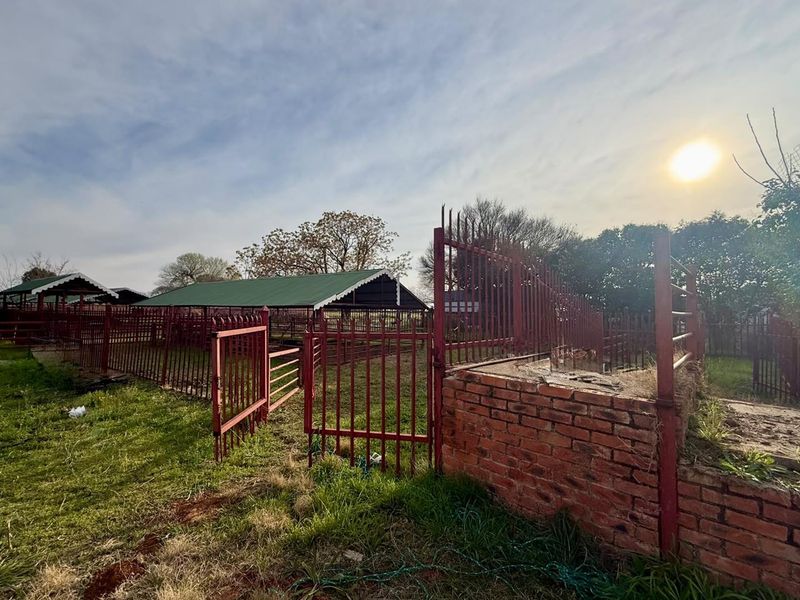 Outdoor farm area featuring metal livestock enclosures, fencing, and a brick structure, set within a rural landscape.