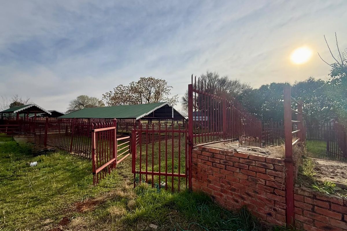 Outdoor farm area featuring metal livestock enclosures, fencing, and a brick structure, set within a rural landscape.