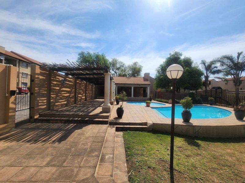 Outdoor pool area featuring a paved patio, a shaded pergola structure with stone columns, and a well-maintained garden setting.