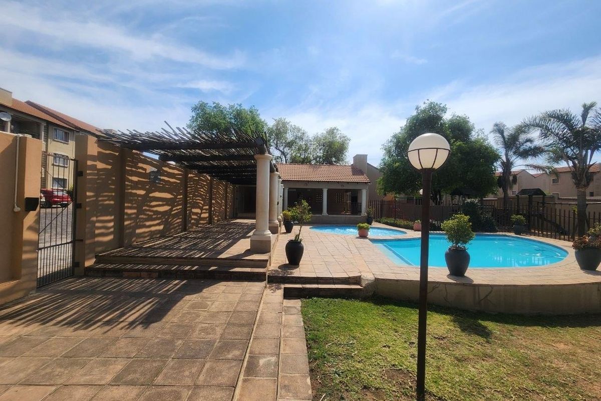 Outdoor pool area featuring a paved patio, a shaded pergola structure with stone columns, and a well-maintained garden setting.