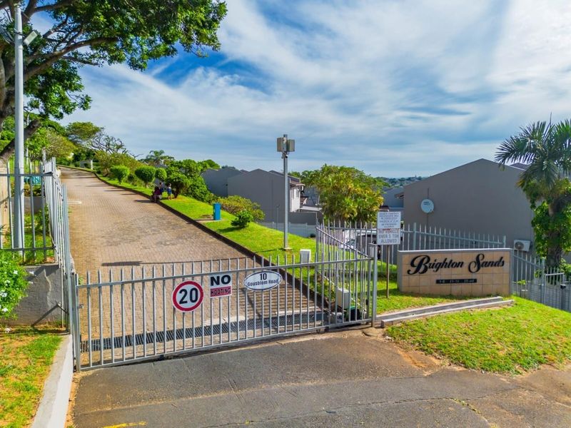 Entrance view of Brighton Sands townhouse complex featuring a secure gated access point, paved driveway, and established landscaping.