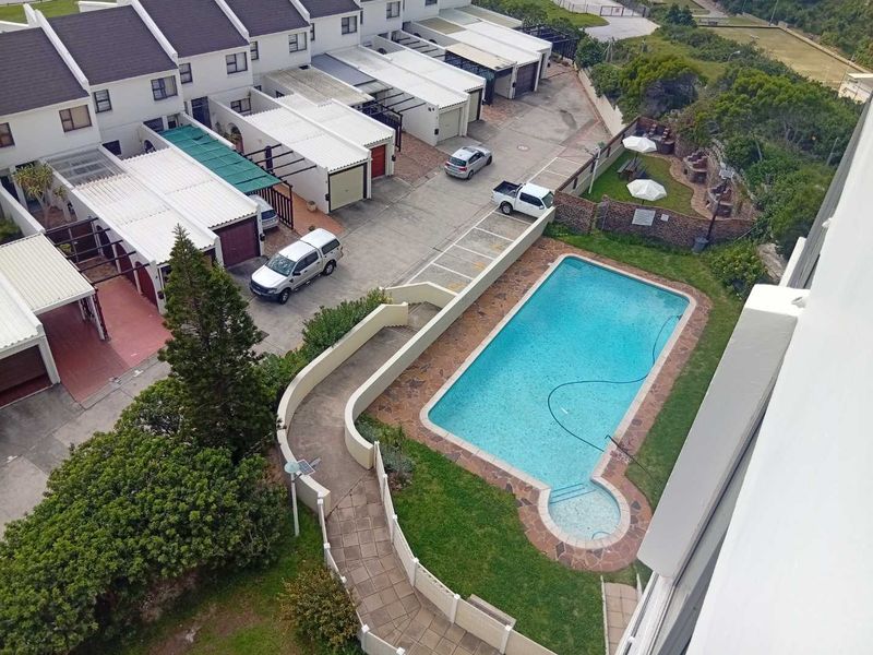 Aerial view of the apartment complex featuring a communal swimming pool, landscaped garden areas, and a paved driveway with garage access.