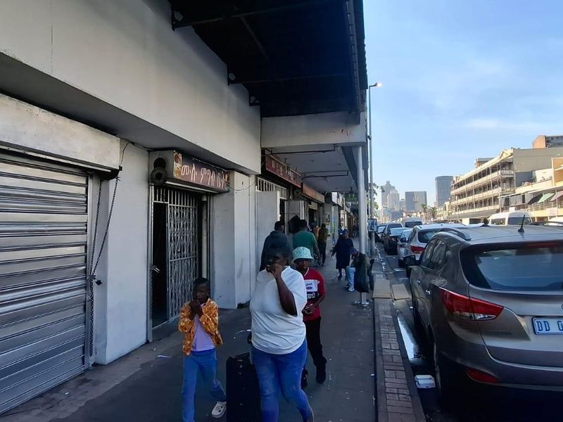 Street-level view of a commercial property featuring metal roller shutter doors and security gates along a busy pedestrian walkway.