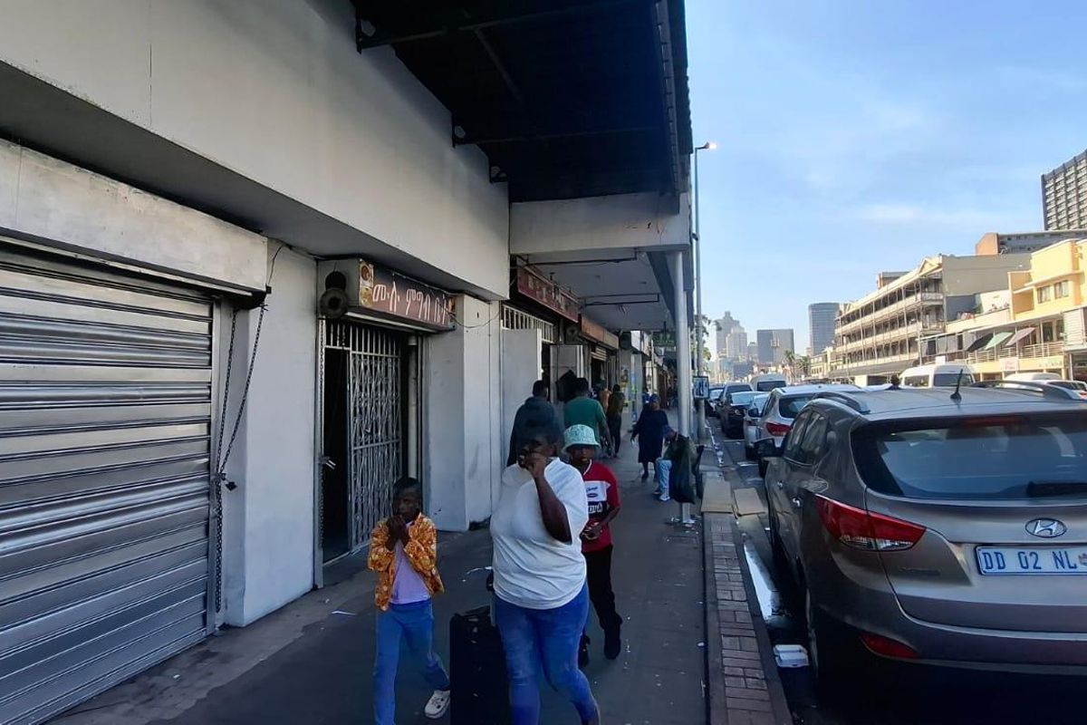 Street-level view of a commercial property featuring metal roller shutter doors and security gates along a busy pedestrian walkway.