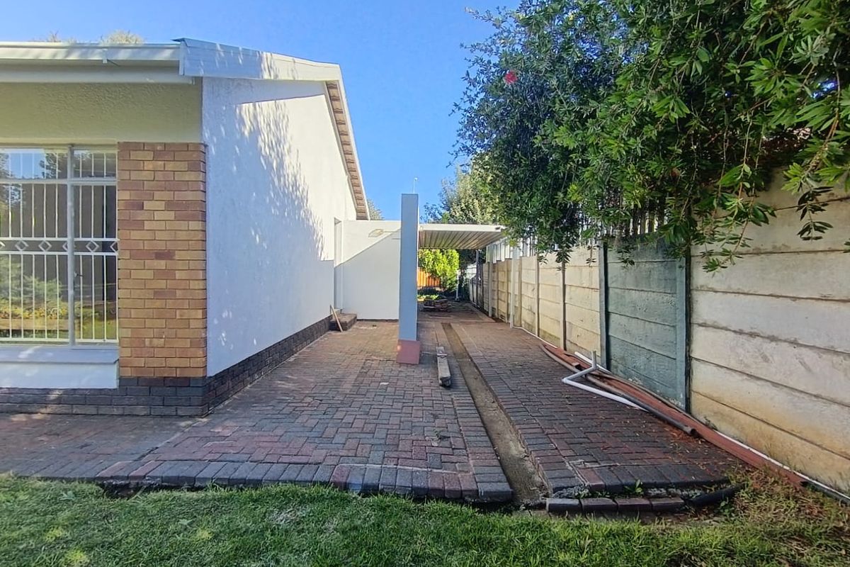 A side exterior view of the property featuring paved pathways, a carport structure, and a perimeter boundary wall.
