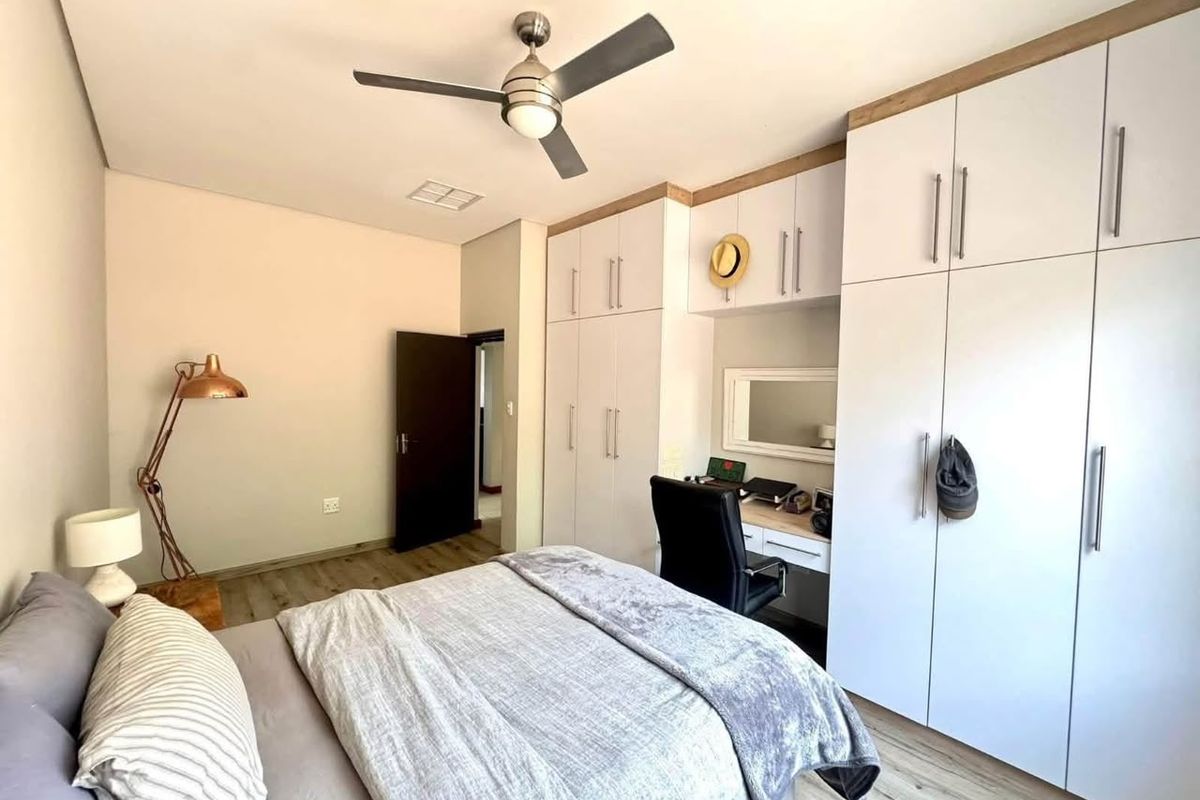 A well-maintained bedroom featuring extensive white built-in cupboards, a integrated desk area, modern laminate flooring, and a functional ceiling fan.