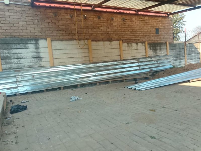 An outdoor area featuring a paved surface, a precast concrete boundary wall, and a brick wall with overhead corrugated metal roofing sheets.