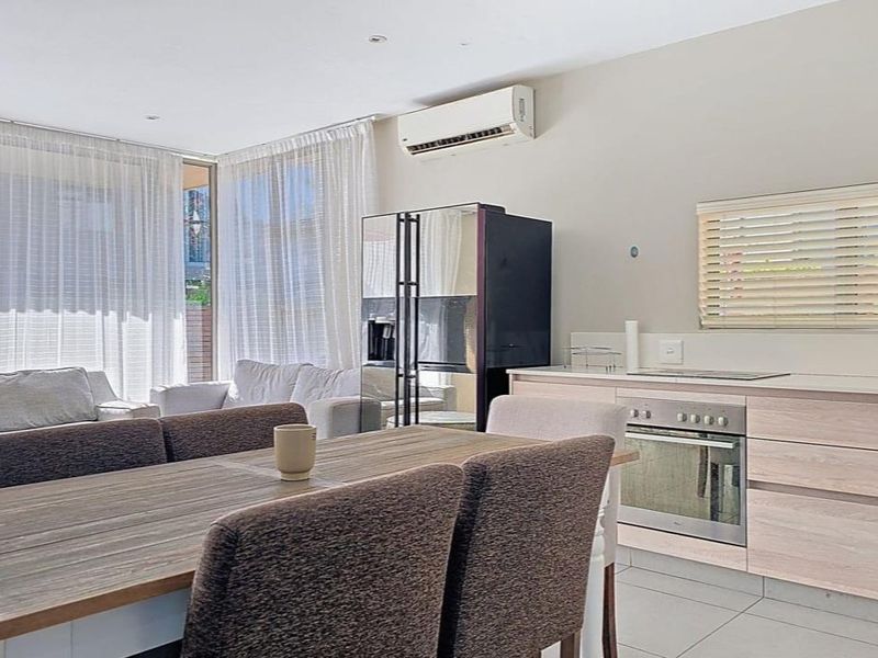 Open-plan living and kitchen area featuring light wood-finish cabinetry, built-in oven, tiled flooring, and a wall-mounted air conditioning unit.