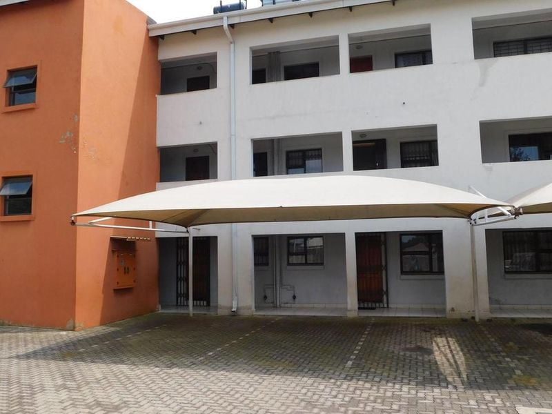 Exterior view of a multi-story townhouse complex featuring a paved driveway with shade sail carports and security-gated entrances.