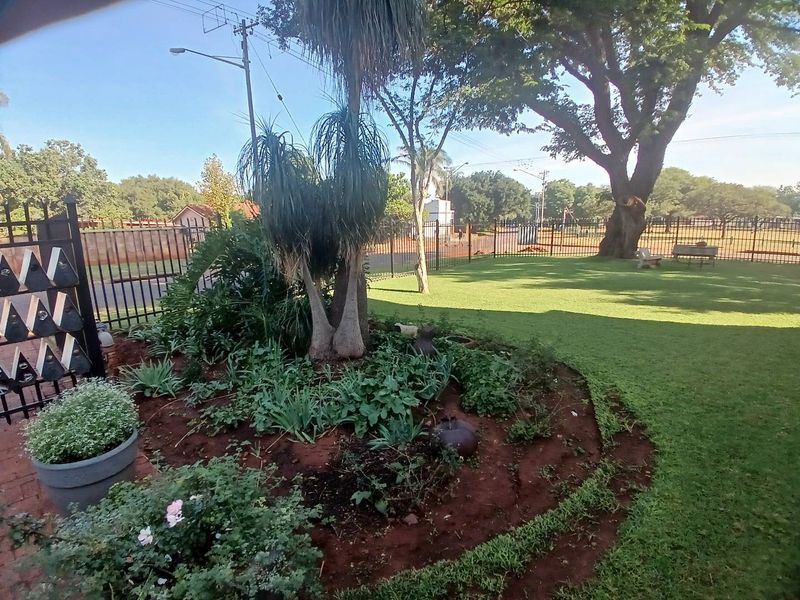 A well-maintained garden area featuring a manicured lawn, mature trees, and established garden beds enclosed by metal perimeter fencing.