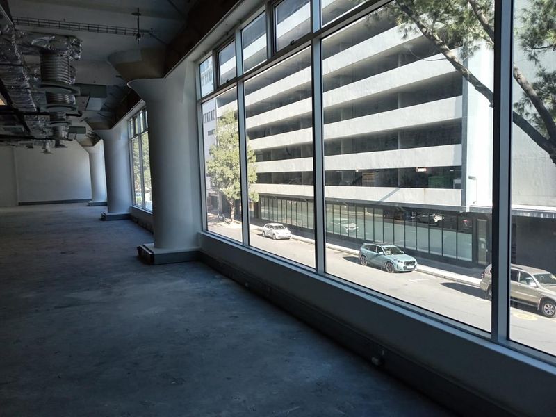 An expansive, empty office space featuring large floor-to-ceiling windows, exposed industrial ceiling ducting, and structural columns with concrete flooring.