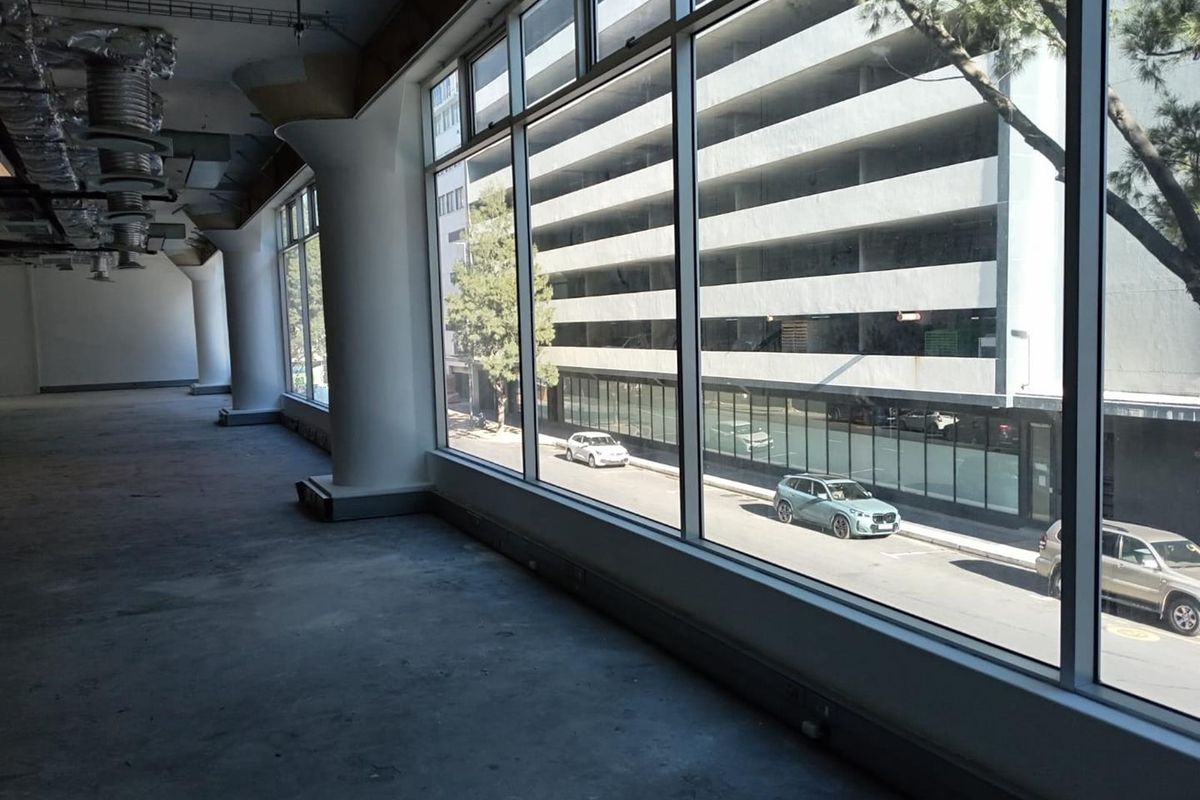 An expansive, empty office space featuring large floor-to-ceiling windows, exposed industrial ceiling ducting, and structural columns with concrete flooring.