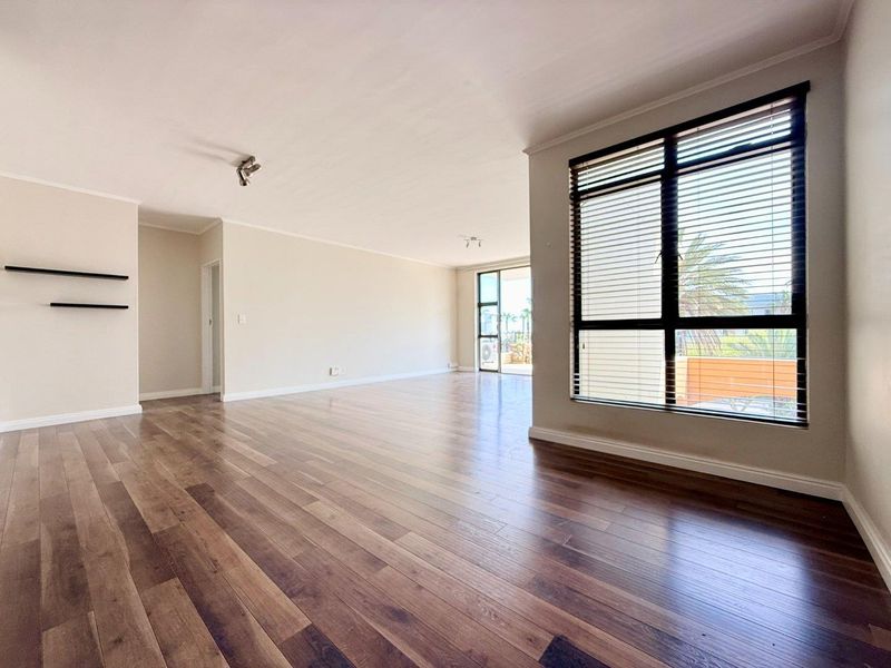 Spacious open-plan living area featuring modern laminate flooring, neutral walls, and large windows that provide ample natural light and access to the balcony.