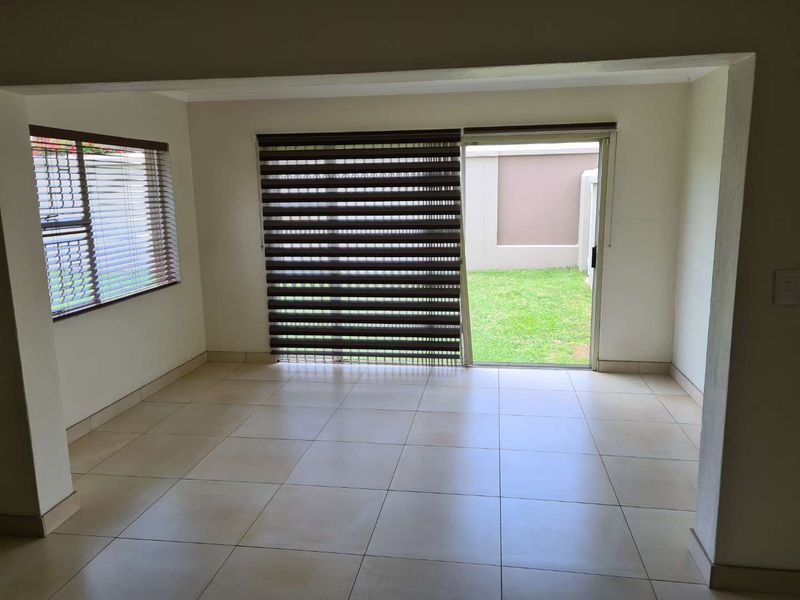 An empty living room featuring neutral tiled flooring, large sliding glass doors leading to the garden, and fitted window blinds.