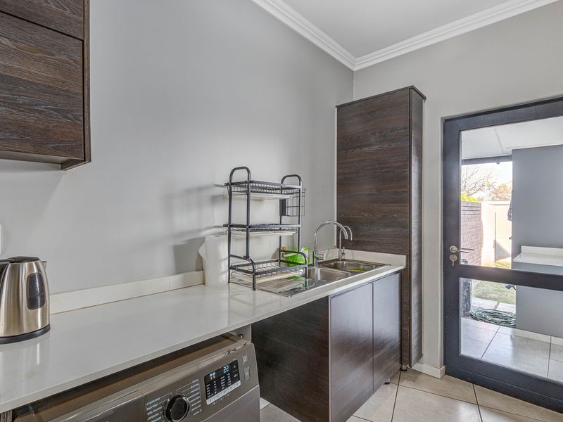 A functional laundry area featuring modern dark wood cabinetry, stone countertops, a stainless steel double sink, and direct access to the exterior.