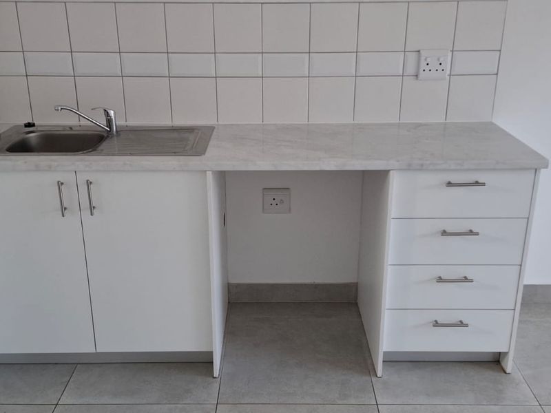 A clean, modern kitchen kitchenette featuring white cabinetry, a stainless steel sink, and a neutral tiled splashback, providing a functional and compact prep area.