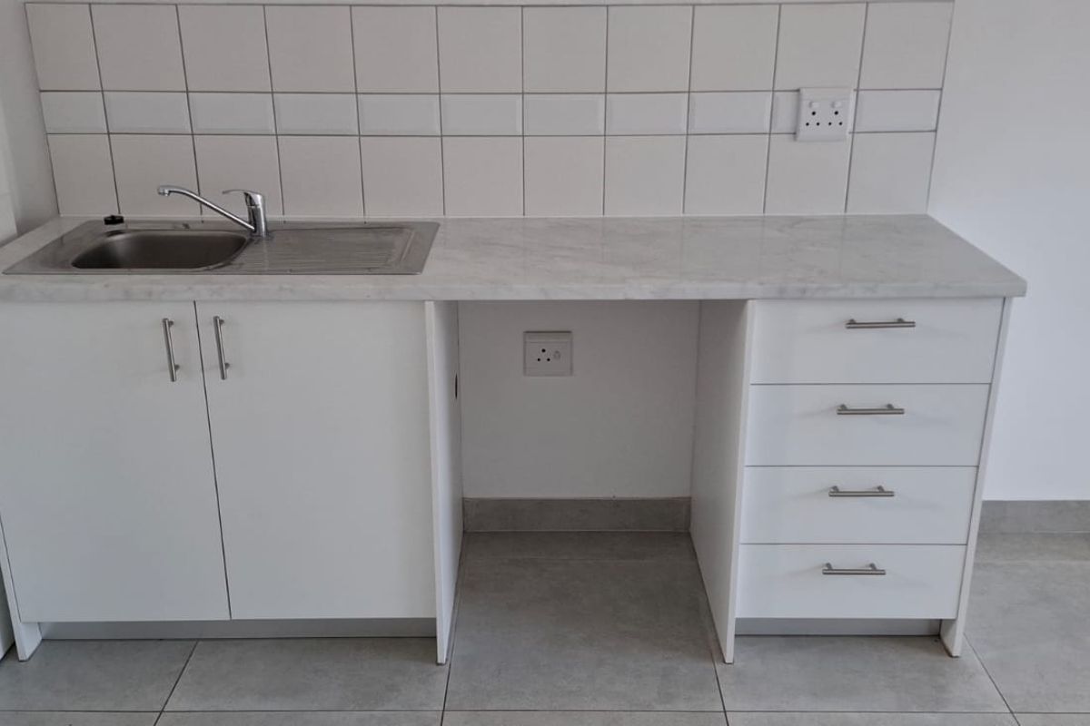 A clean, modern kitchen kitchenette featuring white cabinetry, a stainless steel sink, and a neutral tiled splashback, providing a functional and compact prep area.