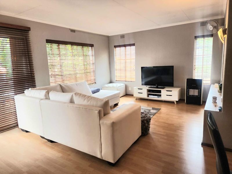 A bright and spacious living room featuring laminate flooring, large windows with venetian blinds, and direct access to an outdoor area via a sliding door.