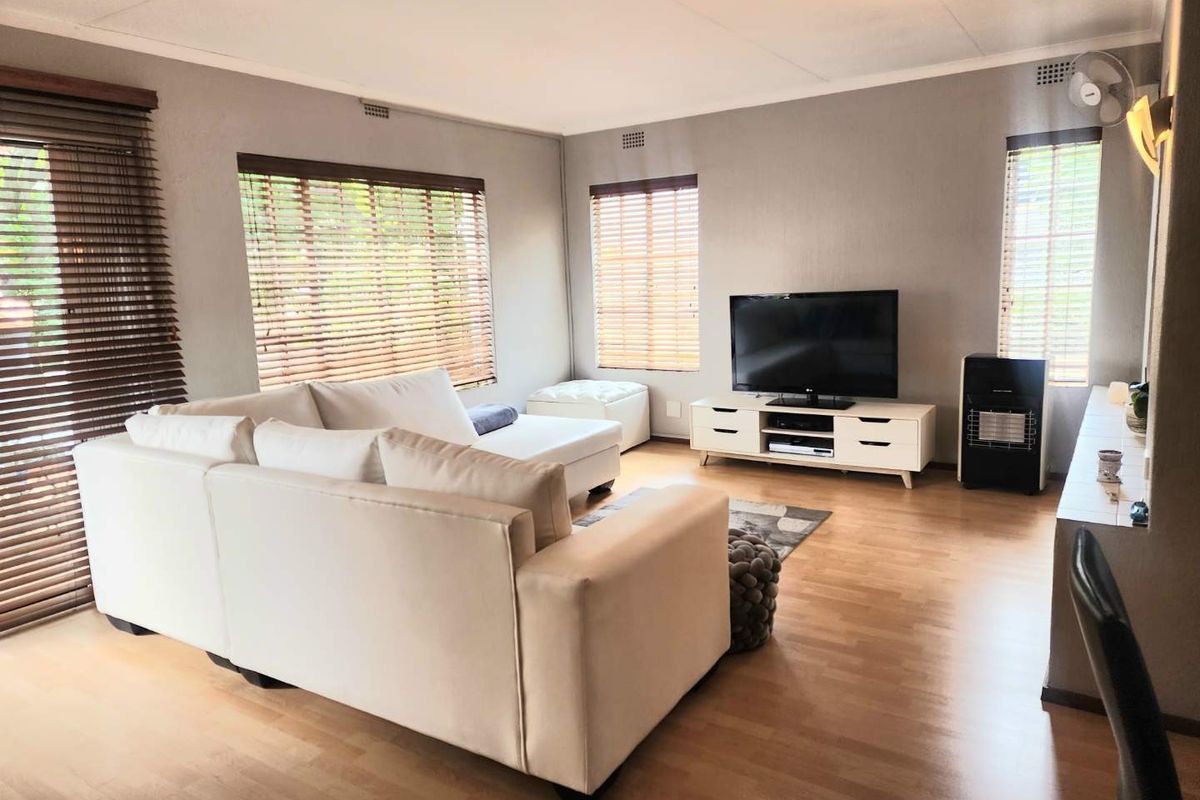 A bright and spacious living room featuring laminate flooring, large windows with venetian blinds, and direct access to an outdoor area via a sliding door.