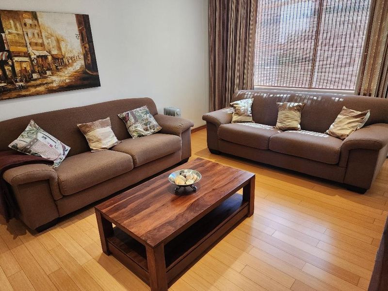 A comfortable living room featuring light-toned laminate flooring and large windows with integrated blinds and curtains.