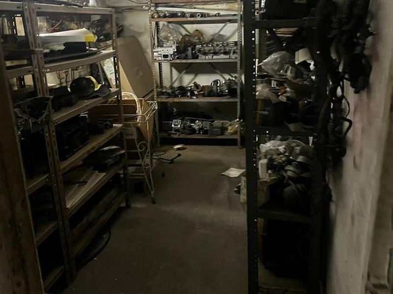 A cluttered utility or storage space featuring concrete flooring and ceiling with exposed electrical wiring, currently used for industrial storage.